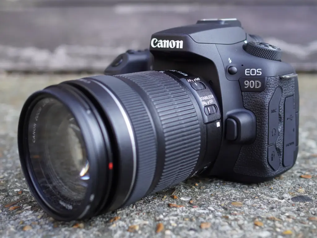 A Canon EOS 90D DSLR camera with a zoom lens is placed on a textured surface.