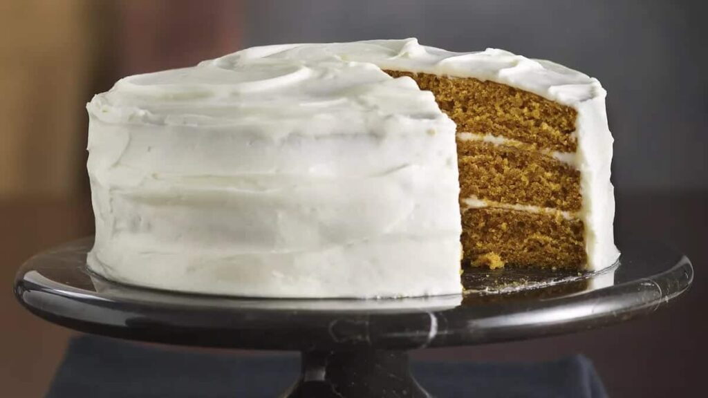 A three-layer cake with white frosting and a slice missing, displayed on a dark cake stand.