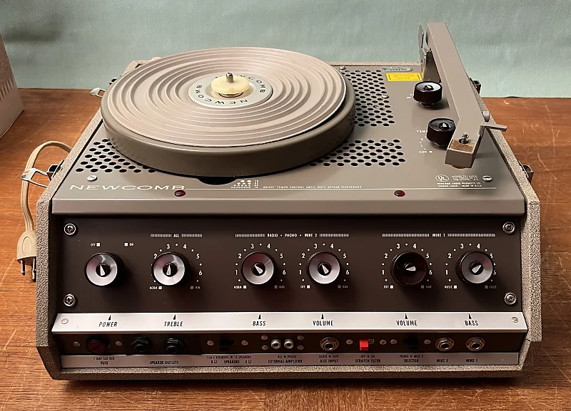 Vintage Newcomb turntable with knob controls for power, treble, bass, and volume, set on a wooden surface.