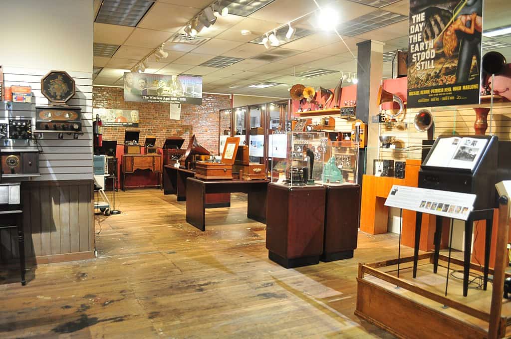 A museum exhibit displays vintage radios, phonographs, and related memorabilia in a well-lit room with wooden floors and informational plaques.