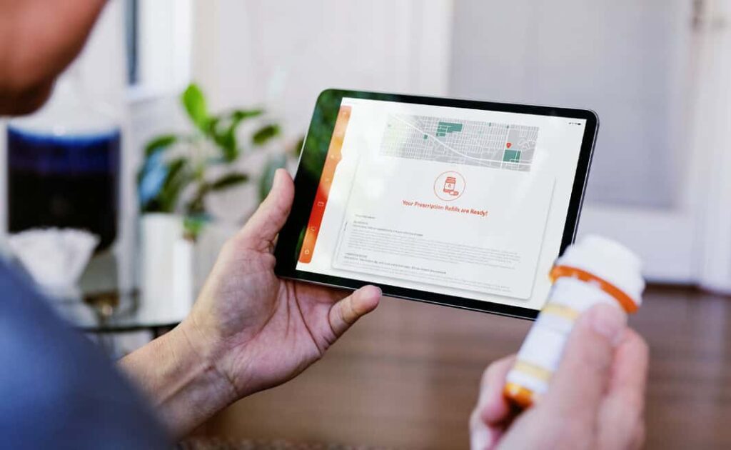 A person holds a tablet displaying a prescription refill notification and a bottle of medication.