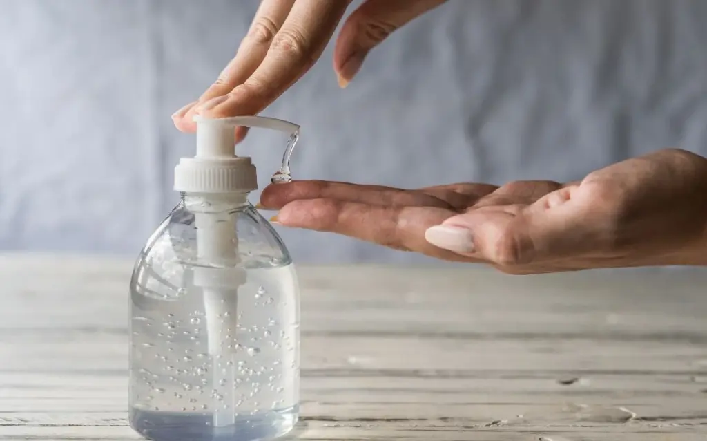 A person dispenses hand sanitizer from a clear pump bottle onto their palm over a white wooden surface.