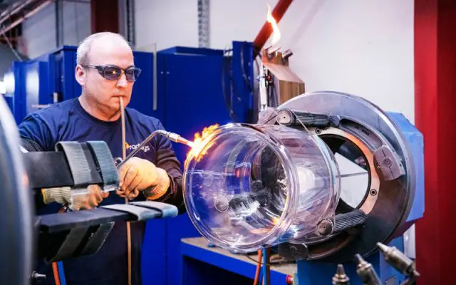 A technician wearing safety glasses uses a torch to work on a large glass cylinder in an industrial setting.