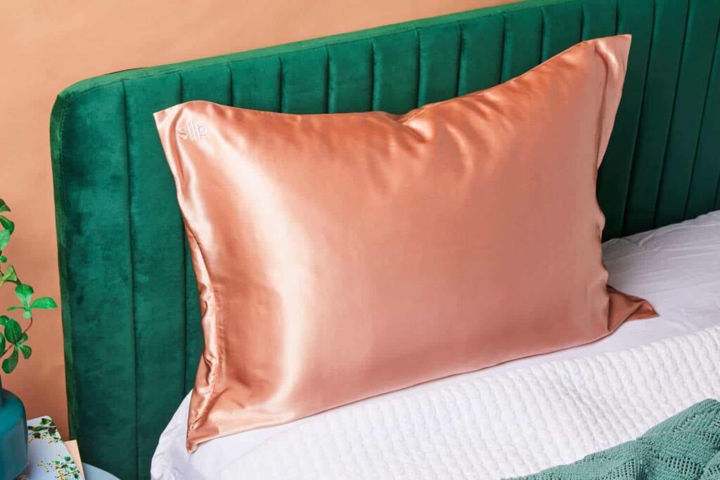 A copper-colored silk pillowcase on a green upholstered headboard, with a white bedsheet and a green knitted blanket.