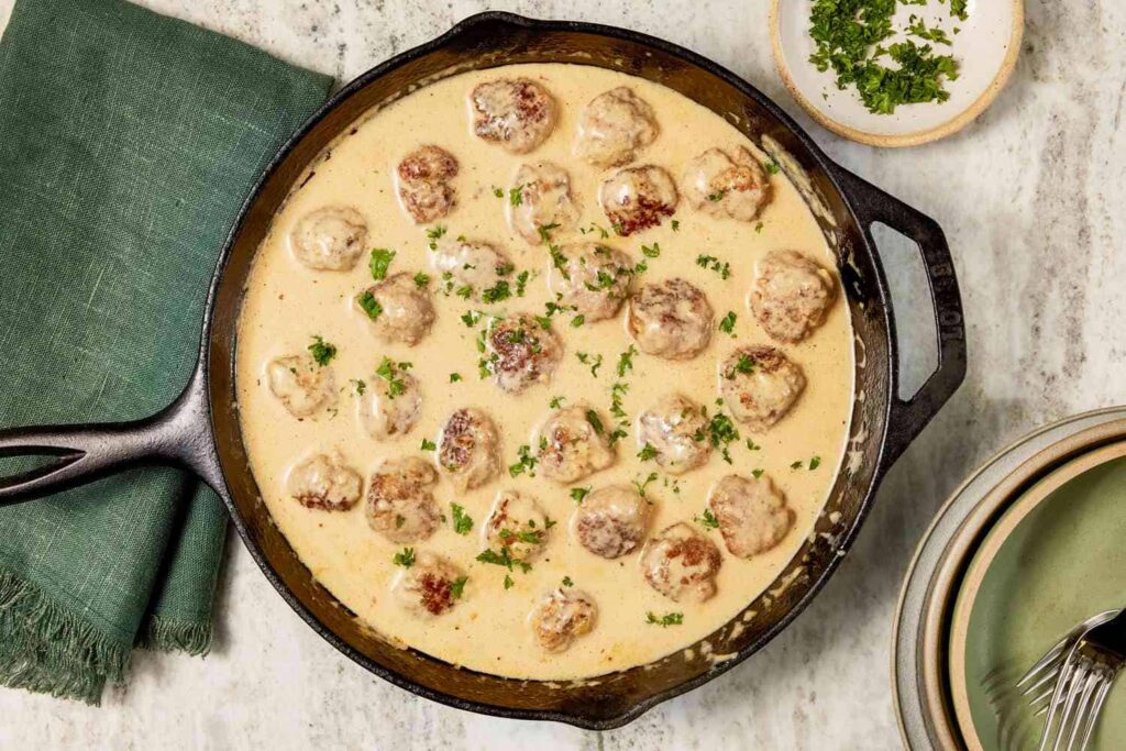 Skillet with meatballs in creamy sauce, garnished with chopped herbs, placed on a marble surface beside a green napkin, small bowl of herbs, and stacked plates.