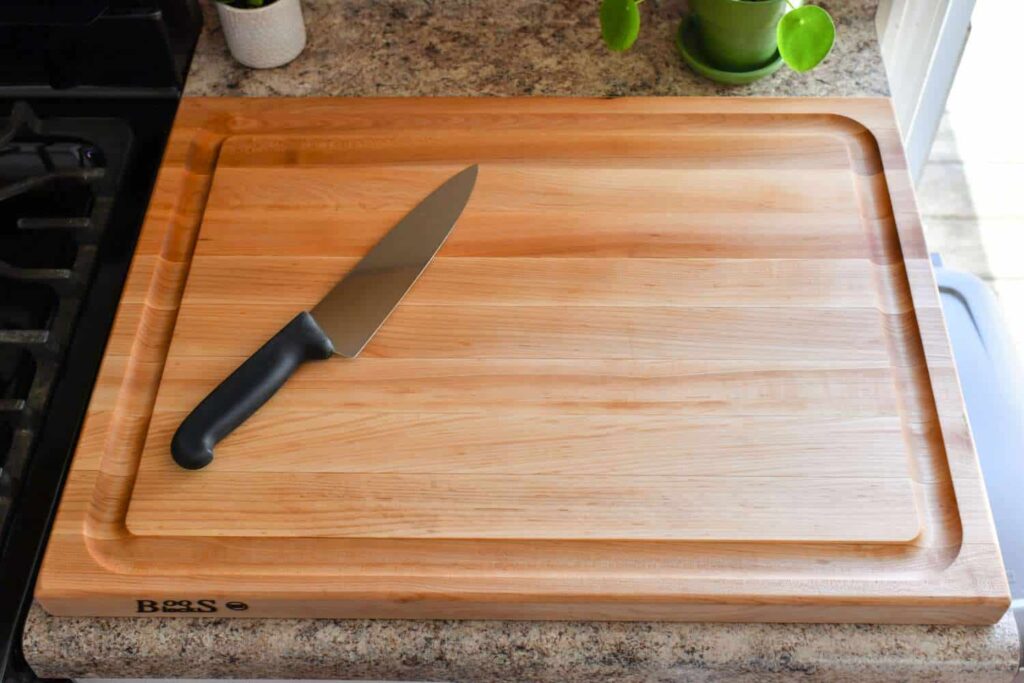 A large wooden cutting board with a grooved edge sits on a countertop. A chef's knife with a black handle rests on the board.