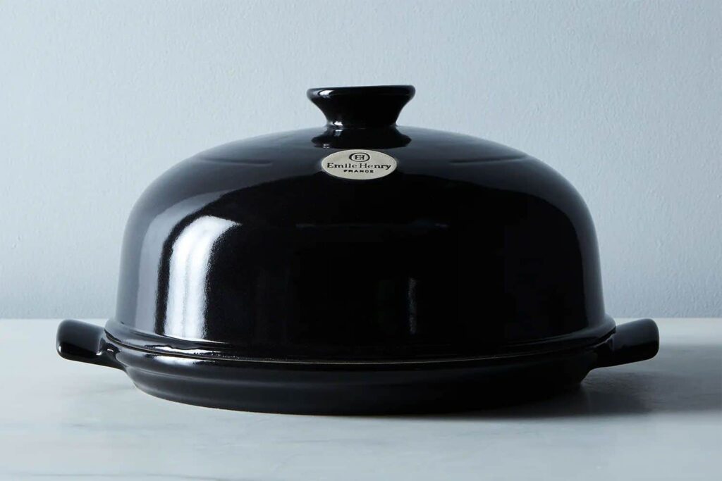 Black ceramic bread cloche with a domed lid, placed on a light-colored surface against a neutral background.