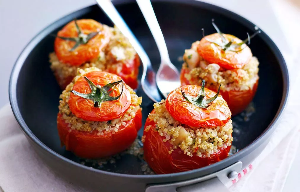 Four stuffed tomatoes filled with quinoa and vegetables in a black pan, each topped with a tomato cap. Two spoons are placed in the pan.