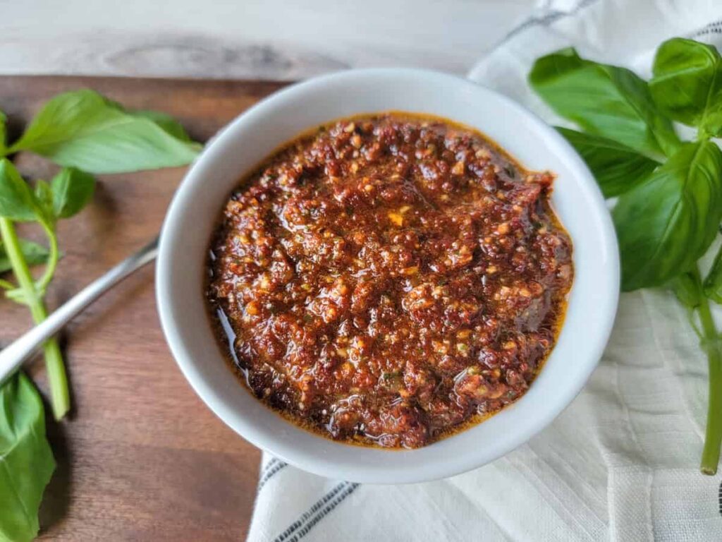 A bowl of red pesto sauce on a white cloth, surrounded by fresh basil leaves on a wooden surface.