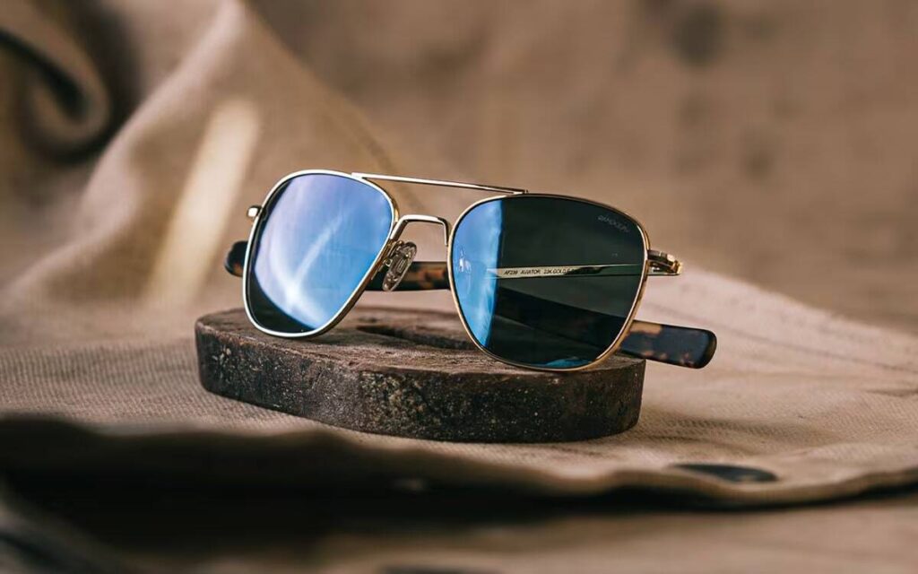 Aviator sunglasses with a gold frame and blue lenses placed on a round wooden block.