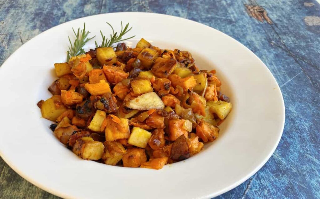 A white plate with a dish of roasted diced potatoes and sweet potatoes garnished with rosemary.