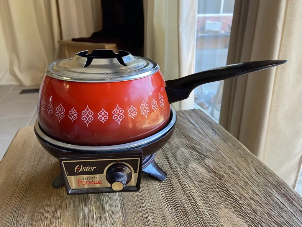 A vintage red electric fondue pot with a black handle sits on a wooden table. It features a decorative white pattern and an Oster brand control dial.