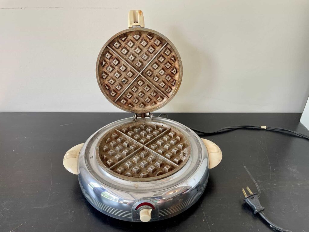 A vintage waffle iron sits open on a black surface, displaying its patterned cooking plates. A power cord is visible to the right.