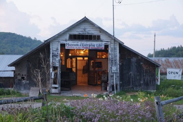 A rustic barn houses the Museum of Everyday Life, with a sign above the entrance. The building is surrounded by grass and flowers, and there is an "OPEN" sign displayed.