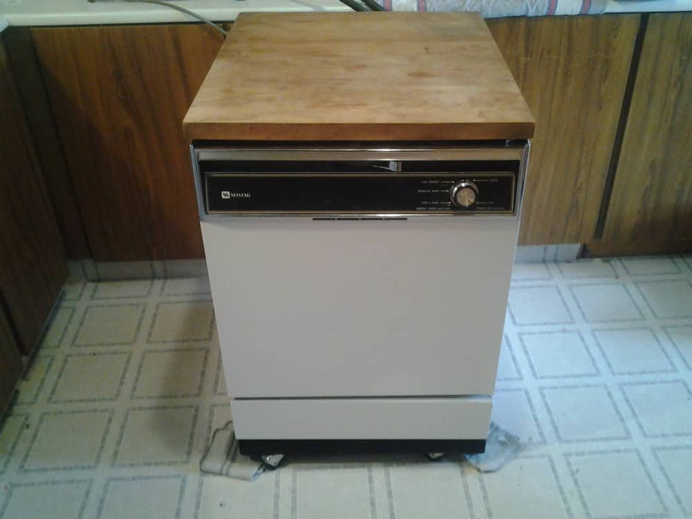 A vintage dishwasher with a wooden top sits on a patterned floor in a kitchen.