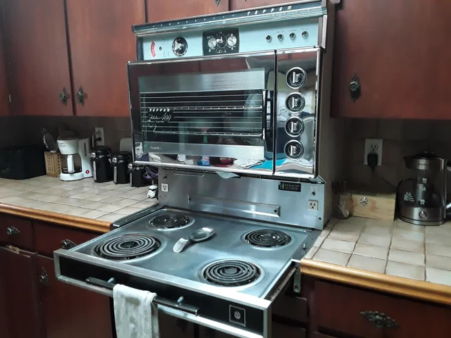 Vintage kitchen with a stainless steel mid-century electric oven and stove, featuring coil burners and a built-in oven, surrounded by wood cabinets and countertop appliances.