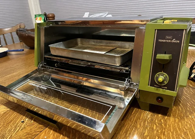 A vintage green toaster oven on a wooden table with the door open, showing a metal tray inside.