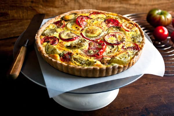 A vegetable quiche with tomatoes and zucchini on a white cake stand, next to a knife. Two tomatoes are in the background on a wire rack.