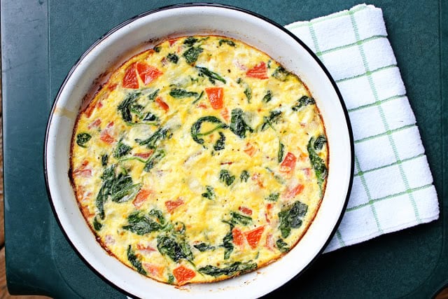 A vegetable frittata in a round baking dish, featuring spinach and tomatoes, rests on a green table next to a folded white and green checkered cloth.