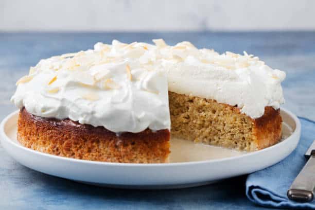 A coconut cake with white frosting and toasted coconut flakes on top, with one slice removed, placed on a white plate.