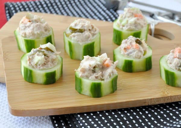 Cucumber slices topped with a tuna salad mixture are arranged on a wooden board with a black and white placemat underneath.