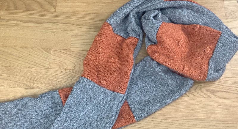 A gray and orange knitted scarf with a textured pattern is laid out on a wooden floor.