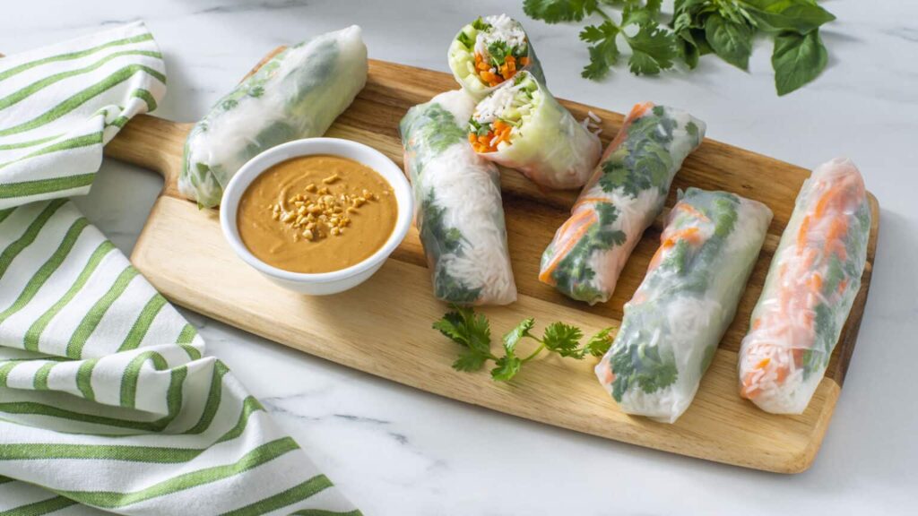 Rice paper rolls with vegetables and herbs on a wooden board, served with a side of peanut dipping sauce. Striped cloth and herbs in the background.