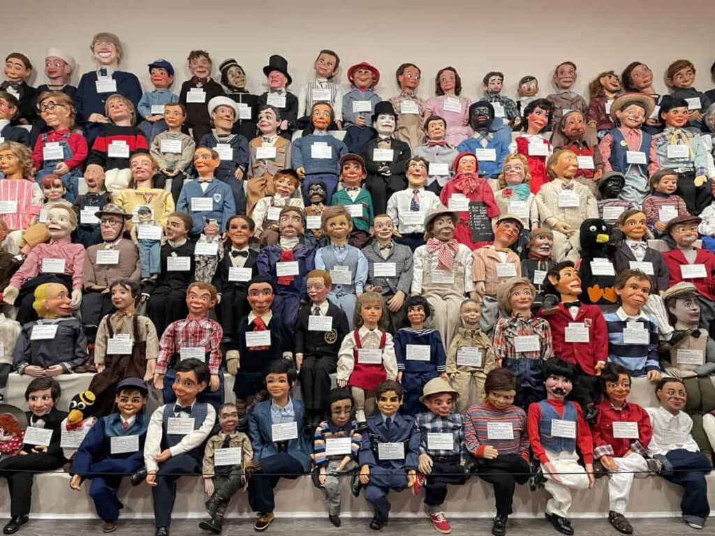 A large collection of ventriloquist dummies is arranged in rows. Each dummy has a name tag, and they display a variety of facial expressions and outfits.
