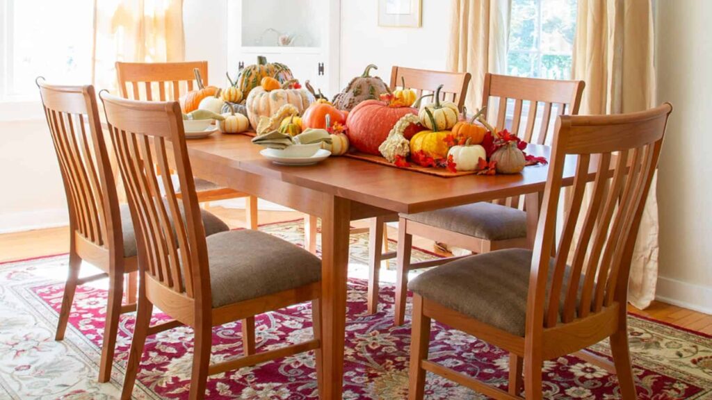 A wooden dining table with six chairs is decorated with an assortment of pumpkins and gourds in a sunlit room.