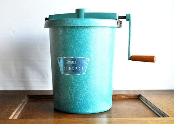 Blue vintage Sears Coldspot ice cream maker with a hand-crank and a wooden handle, on a wooden surface.