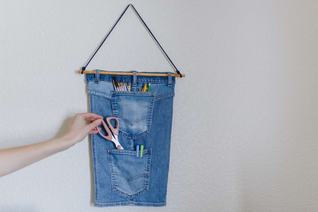Denim wall organizer with multiple pockets holding colorful pencils and scissors, hanging on a white wall.
