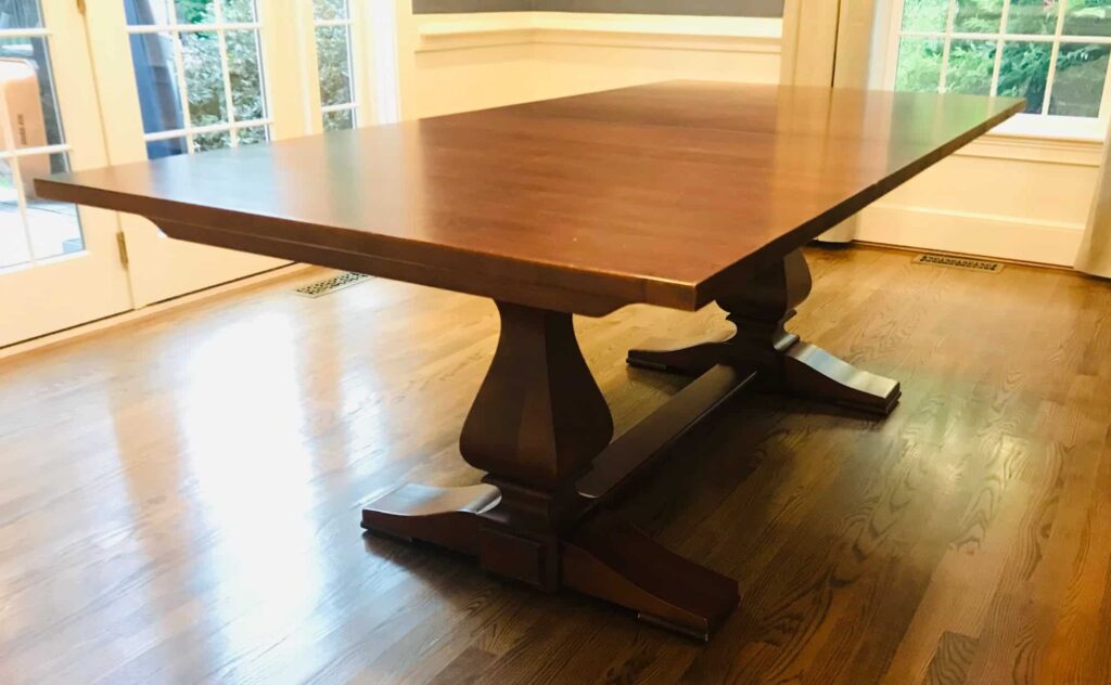 A large wooden dining table with a rectangular top and dual pedestal base sits on a hardwood floor in a bright room.
