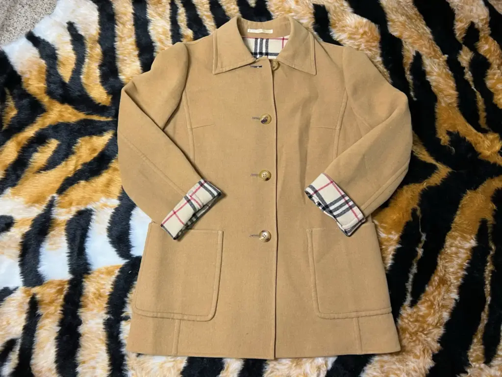 A tan button-up coat with plaid lining on the cuffs is laid flat on a tiger-striped fabric background.