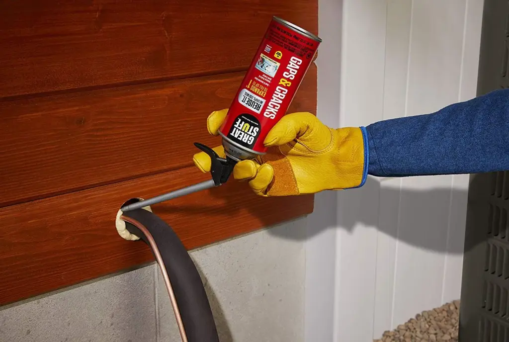 A person wearing yellow gloves uses a can of foam sealant to fill a gap around a pipe entering a wooden wall.