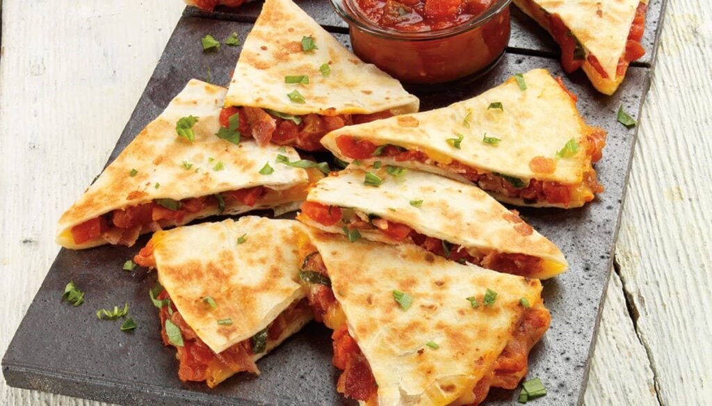 Plate of sliced quesadillas filled with vegetables, topped with chopped herbs, and served with a bowl of salsa.