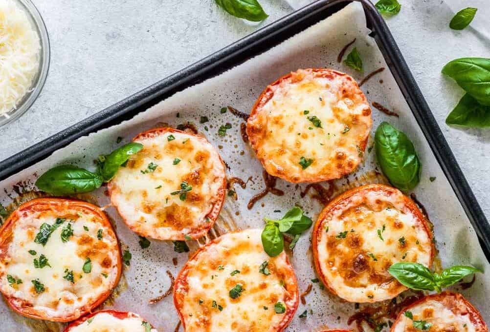 Tomato slices topped with melted cheese and herbs on a baking sheet. Fresh basil leaves are scattered around.