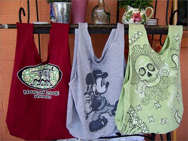 Three tote bags hanging on a shelf: red with "North Shore Hawaii," gray with a cartoon character, and green with a skull design.