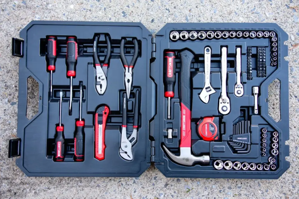 A black toolkit with red and silver tools, including pliers, screwdrivers, adjustable wrenches, a tape measure, and various sockets, is open on a concrete surface.