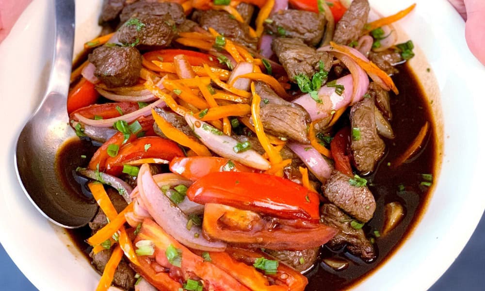 A dish of stir-fried beef, sliced red onions, tomatoes, and carrots in a dark sauce, garnished with chopped herbs, served in a white bowl with a spoon on the side.