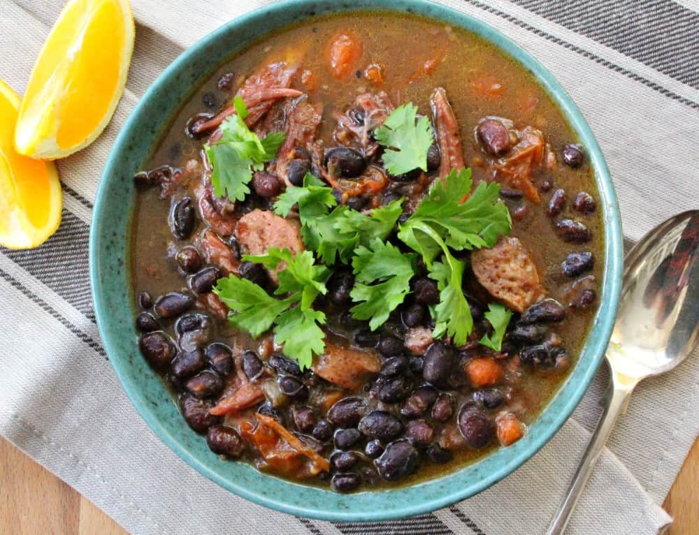 A bowl of black bean stew with meat, garnished with cilantro, placed on a striped napkin next to orange wedges and a spoon.