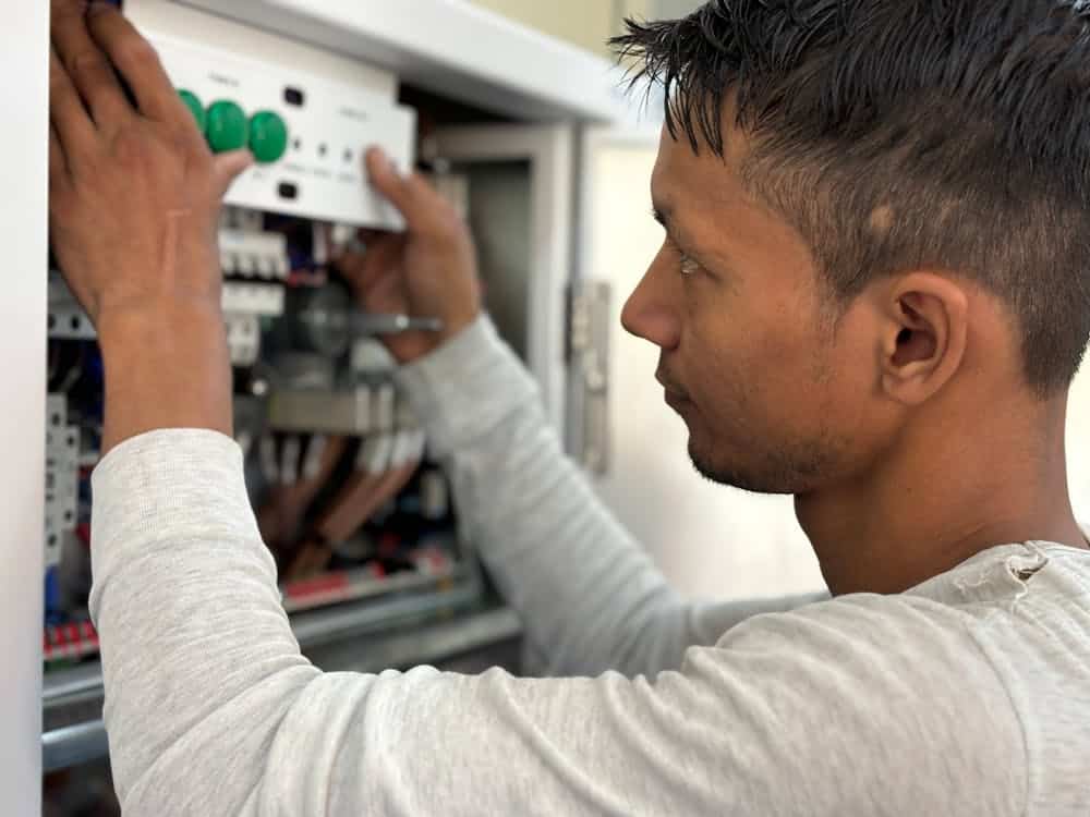 A person in a long-sleeve shirt works on a control panel with wires and components visible.