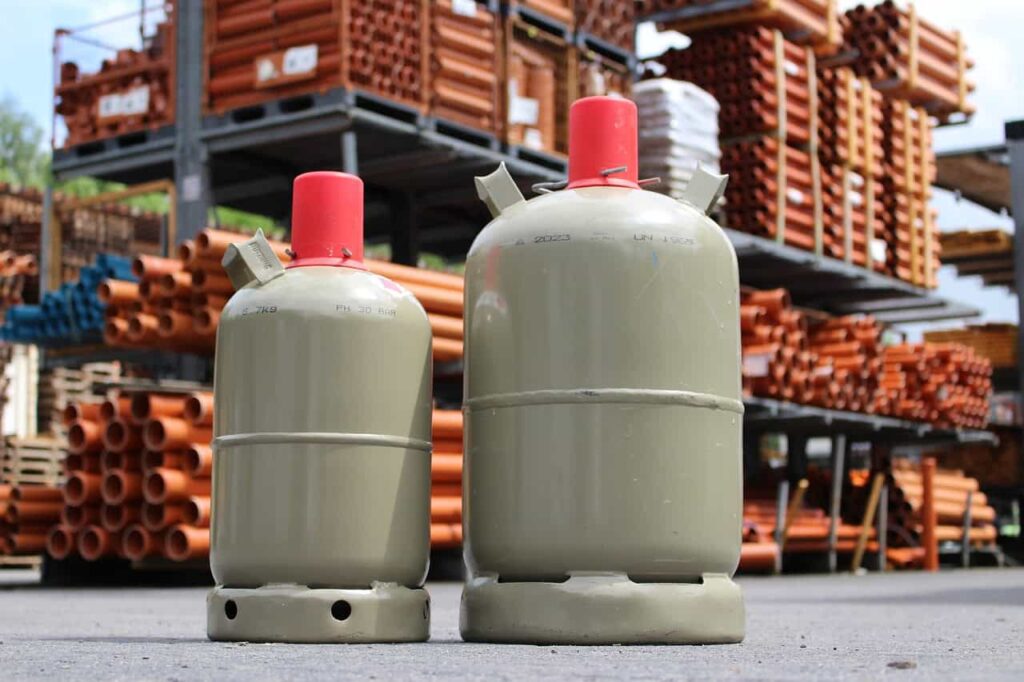 Two large gas cylinders with red caps are placed on the ground in an industrial setting with stacks of pipes in the background.