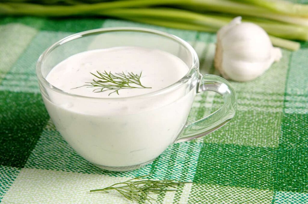 A glass cup filled with creamy white dressing garnished with dill sits on a green checkered cloth, with garlic and green onions in the background.