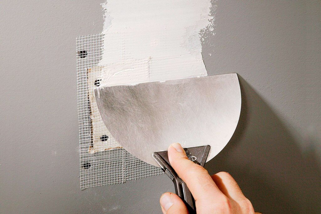 A hand uses a putty knife to apply joint compound over a mesh patch on a gray wall.