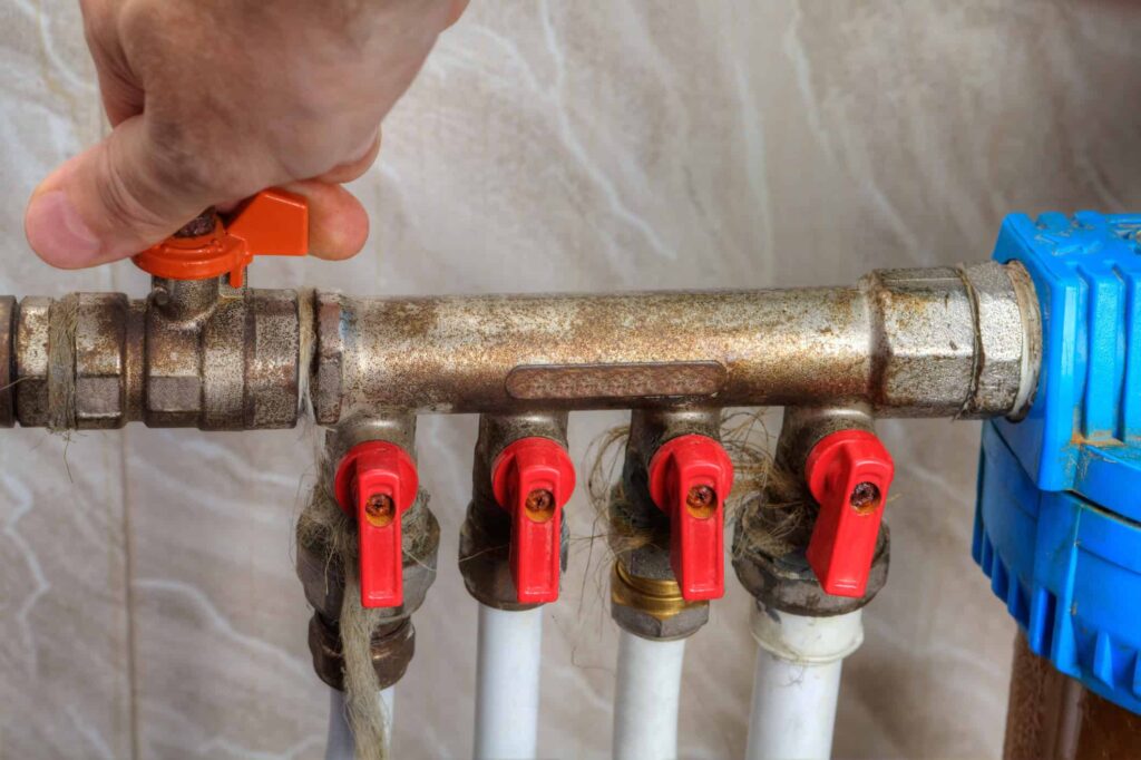 A hand turns an orange valve on a pipe system with four red valves and a blue component.