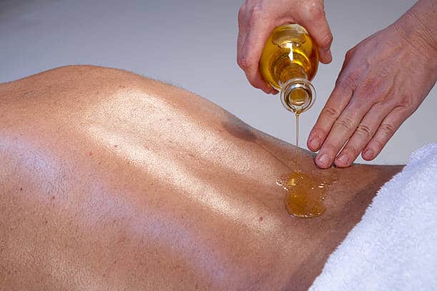 A person pours oil onto another person's bare back in preparation for a massage, with white towels nearby.
