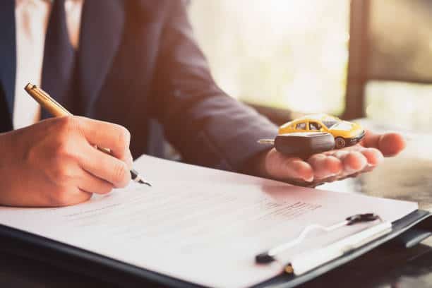 A person in a suit signs a document while holding a toy car and a car key in their hand.