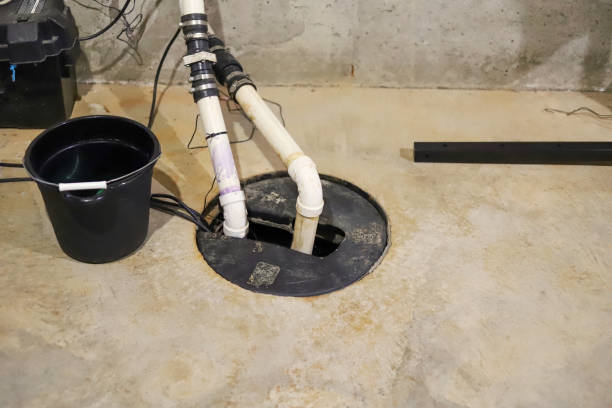 A sump pump system in a basement with PVC pipes and a black bucket on a concrete floor.