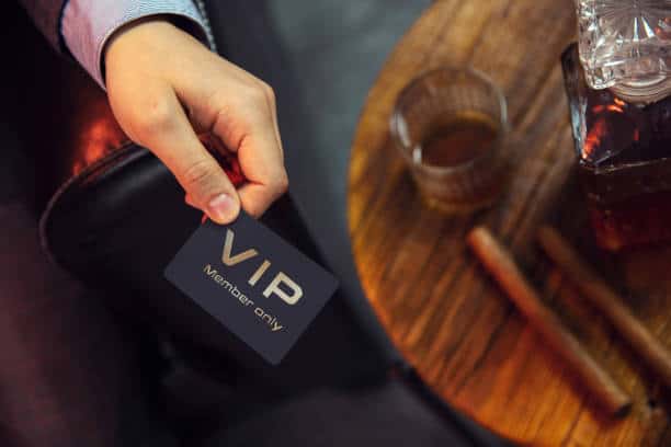 A hand holds a black VIP member card over a wooden table with a glass and a bottle.