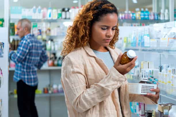 Woman in a pharmacy carefully reads a label on a pill bottle while holding a shopping basket. A person in the background browses the shelves.